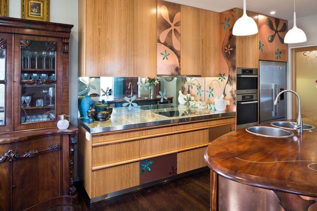 Eclectic, richly detailed kitchen with warm timber-grain cabinetry, dark timber flooring, a reflective patterned glass splashback, dark stone benchtops, integrated stainless-steel appliances, and a distinctive rounded timber island with a double-bowl sink. Decorative floral motifs and a traditional dark-wood display cabinet add a vintage character to the space, while white pendant lights provide contrast.