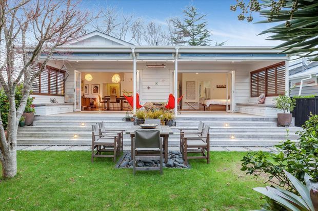 Wide view of a refined single-storey home opening onto a landscaped lawn and stepped terrace. The white weatherboard facade has a shallow pitched gable roof, extensive open glazing or doors, timber louvred shutters, built-in white seating, and a central outdoor dining and lounge arrangement. Broad pale stone or concrete steps feature integrated lighting, with mature planting framing the scene. Interior living and dining areas are visible through the open frontage.