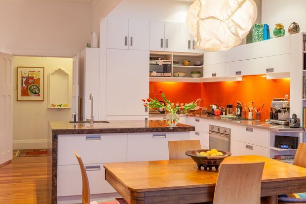 A bright, warm kitchen and dining area with extensive white built-in cabinetry, a dark stone island benchtop, vivid orange-red glass or glossy splashback, stainless-steel appliances, and timber dining furniture. The kitchen opens toward a hallway or adjoining room, with decorative display shelving and a large sculptural pendant light.