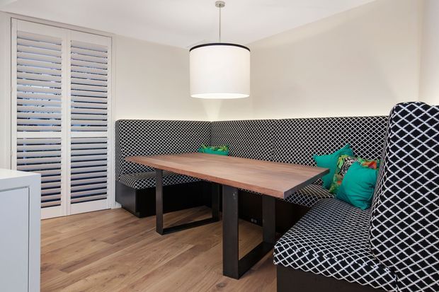 A compact dining nook with a built-in L-shaped upholstered banquette, upholstered in a bold black-and-white diamond pattern. A rectangular timber dining table with dark trestle-style legs sits in front of the seating, accompanied by bright teal and patterned cushions. The space has light neutral walls, timber-look flooring, plantation shutters, and a simple cylindrical pendant light.