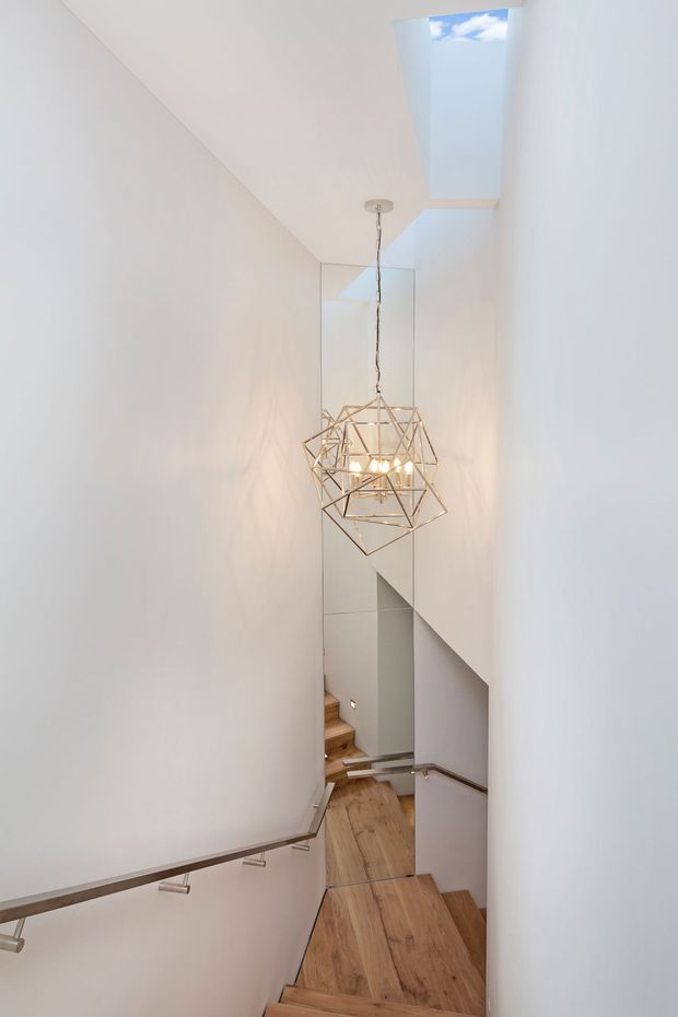 Narrow, minimalist stairway descending through a tall white-painted stairwell. The space features warm timber stair treads and flooring, slim metal handrails, a full-height mirrored wall, a geometric brass-toned pendant light, and a long roof skylight bringing in daylight.
