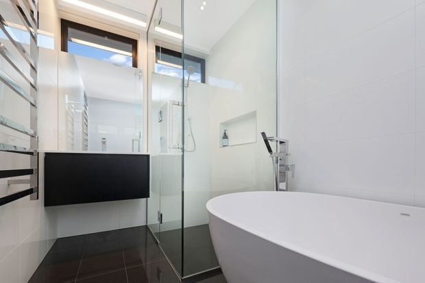 Contemporary bathroom with a freestanding white bathtub, frameless glass shower enclosure, wall-mounted black vanity, recessed shower niche, chrome fixtures, large white wall tiles, and dark tiled flooring. A mirrored cabinet and bright clerestory glazing add light and reflect the compact, highly finished space.
