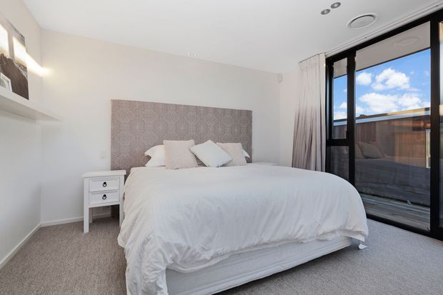 Bright, contemporary bedroom with a white upholstered bed dressed in white bedding, a patterned padded feature headboard, white bedside table, light carpet, and large black-framed glazing with curtains. The room has a clean neutral palette and opens visually toward an exterior balcony or terrace.