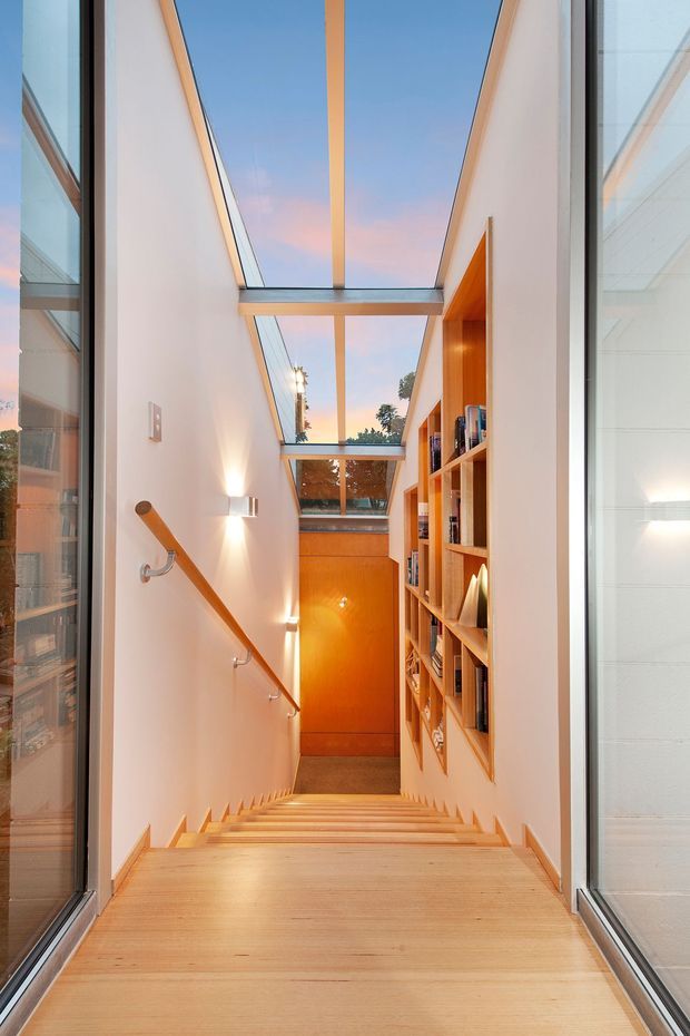 A narrow, bright interior stairway descends between white walls, with a light timber staircase, timber handrail, and built-in timber bookshelf along the right wall. Large glazed openings and overhead skylight-like roof glazing bring in daylight and reveal the blue evening sky. Warm wood and orange-toned joinery contrast with the clean white surfaces, creating a modern, airy circulation space.