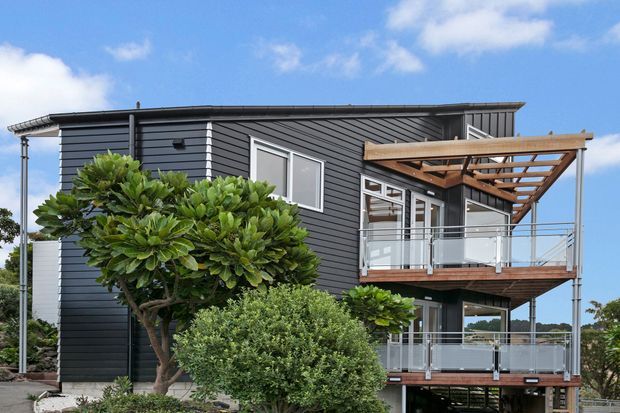 A modern multi-level house with dark horizontal cladding, a low-pitched flat roof, extensive glazing, and two elevated balconies. A timber pergola frames the upper balcony, while landscaped shrubs and sculpted trees surround the exterior. The image is photographed from outside under a bright blue sky.