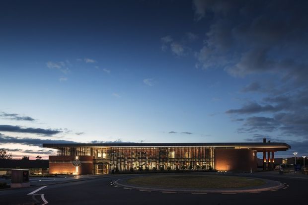 A wide dusk exterior view of a large contemporary civic or commercial building. The low, horizontally composed structure has a projecting flat roof, extensive illuminated floor-to-ceiling glazing screened by vertical fins, and warm brick or terracotta-coloured solid volumes at either end. It fronts a curved paved drop-off drive and landscaped forecourt beneath a dramatic blue evening sky.