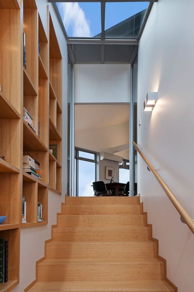 A modern, light-filled stairway with broad timber-clad steps, integrated timber shelving along the left wall, and a simple timber handrail on the right. A glazed roof section and tall windows bring in daylight, with a dining or living area visible at the upper landing.