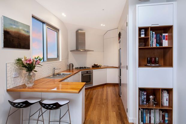 A bright, compact contemporary kitchen with white flat-panel cabinetry, warm timber flooring and a timber-topped peninsula with two bar stools. The kitchen includes stainless steel appliances, a tiled textured splashback, a gas cooktop, undermount sink and integrated storage. A built-in timber bookshelf is visible at the right, while a window brings in natural light.