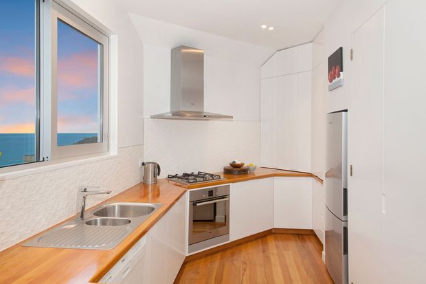 A bright, compact contemporary kitchen with a U-shaped layout, white flat-panel cabinetry, warm timber benchtops and flooring, stainless-steel sink and appliances, a textured white splashback, and a large ocean-view window.
