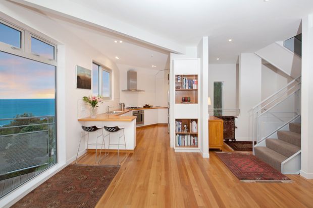 Bright open-plan interior combining a compact kitchen, breakfast bar, living or entry area, and staircase. The space features warm timber flooring, white walls and cabinetry, a built-in white bookcase, patterned rugs, and large glazed openings with an ocean view. The staircase has carpeted treads and a simple white balustrade.