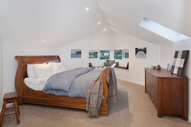 Bright, spacious attic-style bedroom with a pitched white ceiling, recessed lighting, and a skylight. The room features a traditional dark wood sleigh bed, matching wooden dresser, neutral carpet, soft white bedding, and muted blue-grey throws. Built-in window seating and a row of windows overlook greenery, while simple artwork and a small stool complete the calm, refined interior.