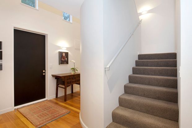 Bright residential entryway and stairwell with a tall white curved or rounded wall enclosing a carpeted staircase. The entry features a matte black door, warm timber flooring, a patterned area rug, a traditional wooden console table, and high clerestory windows. White walls, simple wall lighting, and a white stair handrail create a clean, light-filled interior.