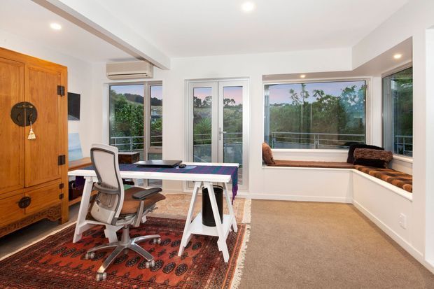 Bright home office with a large wooden storage cabinet, modern desk and ergonomic office chair, patterned area rug, beige carpet, and built-in window seating overlooking greenery. White walls, abundant glazing, and a compact built-in workspace create an airy, functional room.