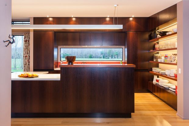 A sophisticated contemporary kitchen featuring dark timber cabinetry, a large central island with a warm-toned benchtop, integrated open shelving displaying glassware and crockery, and a long horizontal window above the rear counter. Light timber flooring and warm layered lighting create contrast against the dark joinery.
