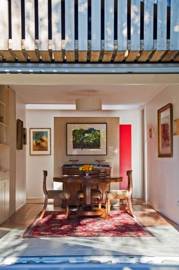 A compact, refined dining room viewed through a wide opening beneath an overhead slatted structure. The room features a dark wood dining table, upholstered dining chairs, a timber sideboard, framed artwork, a patterned red rug, and a vivid red accent wall or doorway. Warm daylight enters from the foreground, while the upper portion shows timber and metal slat detailing.
