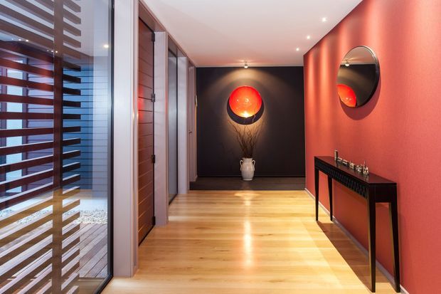 A polished contemporary residential hallway or entry corridor with warm timber flooring, dark charcoal feature wall, and a vivid terracotta-red wall with a round mirror. A slim black console table sits beneath the mirror, while a glowing red spherical wall light and a decorative vase create a dramatic focal point. Full-height glazing and horizontal timber screening appear along the left side, with recessed ceiling spotlights and timber-framed doors adding architectural detail.
