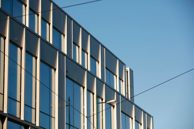 Close-up exterior view of a contemporary multi-storey building facade with a strong geometric grid of tall blue-grey glazed panels, dark metal framing, and projecting vertical fins. Overhead utility wires cross the clear sky, while warm sunlight creates alternating highlights and shadows across the facade.