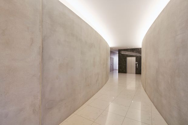 A minimalist interior hallway with gently curving plaster or concrete-finished walls, a glossy pale tiled floor, and concealed linear ceiling lighting. The corridor leads toward a dark stone feature wall and a simple light-coloured door, creating a refined, gallery-like atmosphere.