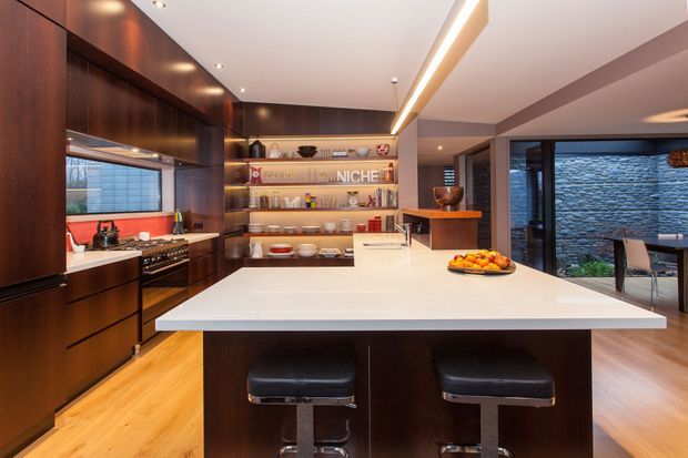 Contemporary open-plan kitchen with dark timber cabinetry, a large white island with two stools, integrated linear ceiling lighting, open shelving, stainless-steel appliances, and glazing opening toward a stone-clad outdoor area.