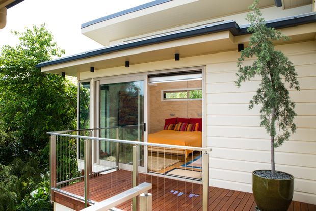 Exterior view of a contemporary upper-level deck and addition with horizontal light-coloured cladding, broad layered roof eaves, sliding glass doors, and slim metal balustrades. The deck is furnished as a compact outdoor sitting area and opens into a bright bedroom or living space with a vivid orange bed or sofa visible inside. Lush planting surrounds the elevated timber deck.