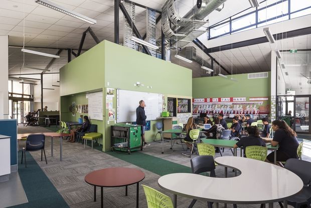 A bright contemporary educational common area or classroom with an open-plan layout, green feature walls, multiple round tables, colorful chairs, visible student work, and a teacher or presentation area. The space has an exposed industrial-style ceiling with ductwork, suspended linear lighting, structural framing, and extensive clerestory glazing.