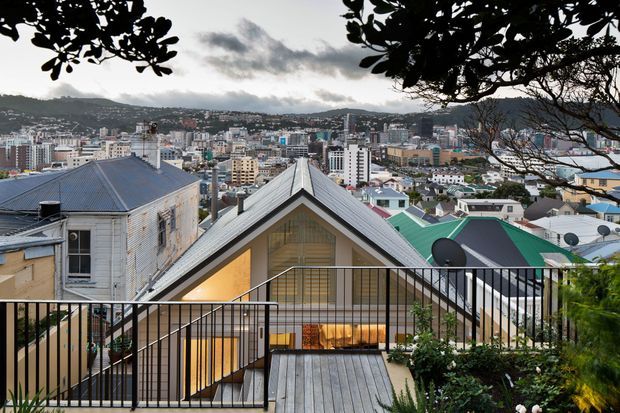 Elevated exterior view from a landscaped terrace or balcony overlooking a densely built hilly urban neighbourhood. The foreground shows a contemporary gabled dwelling with silver metal roofing, pale cladding, extensive glazing, black metal balustrades, timber decking and warm interior lighting, framed by mature foliage. Numerous surrounding rooftops and apartment buildings fill the middle ground, with hills in the distance.