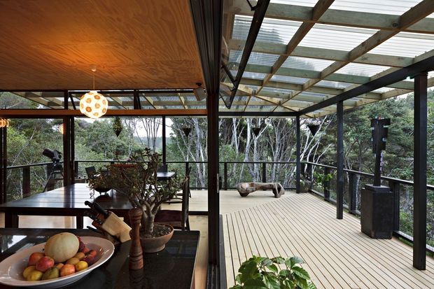 A contemporary enclosed dining and living space opens onto a large timber deck through extensive dark-framed glazing. The deck is sheltered by a translucent pergola roof supported by exposed dark structural beams, while the interior features a timber ceiling, dining table, pendant light, plants, and a kitchen counter with fruit bowl. Dense vegetation surrounds the elevated-looking outdoor platform.