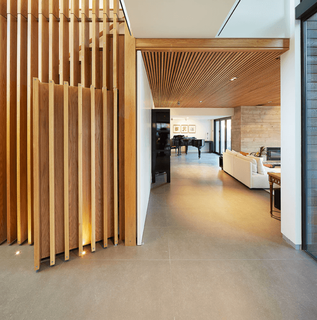 A contemporary open-plan interior viewed from an entry or circulation area, featuring a dramatic vertical timber batten screen with integrated low lighting, a matching slatted timber ceiling, polished grey concrete flooring, white walls, and a spacious living and dining area beyond. Large glazed openings provide natural light, while a white sofa, dining table, framed artwork, and dark joinery furnish the background.