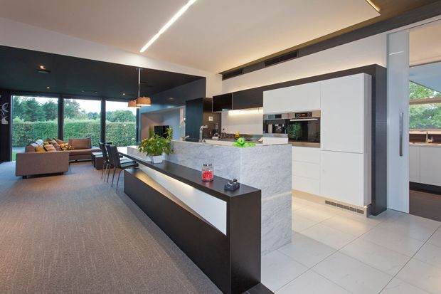 Open-plan contemporary kitchen and living area with a large white marble-look island, streamlined white cabinetry, dark ceiling and joinery accents, integrated appliances, and a long black island extension with seating. Floor-to-ceiling glazing opens the living area toward greenery outside, while recessed linear lighting creates a clean modern atmosphere.