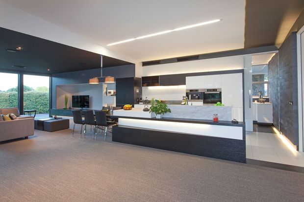 Open-plan contemporary kitchen and living area with a large white island, dark charcoal cabinetry, integrated appliances, illuminated joinery, and a textured stone feature wall. Floor-to-ceiling glazing opens toward greenery, while a dining and lounge area sits adjacent to the kitchen.
