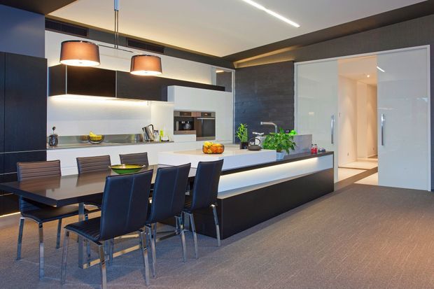 Contemporary open-plan kitchen and dining area with a long dark island, integrated white cabinetry, stainless-steel appliances, pendant lighting, and a six-seat dining table. A textured dark feature wall and recessed linear lighting add contrast, while a wide doorway leads to an adjacent hall or room.