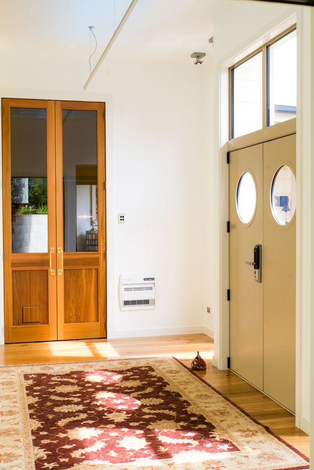 Bright residential entryway with warm timber glazed double doors, a contrasting pale front door with circular windows, white walls, timber flooring, and a large patterned rug. Natural light enters through the glazed entry doors and side windows.