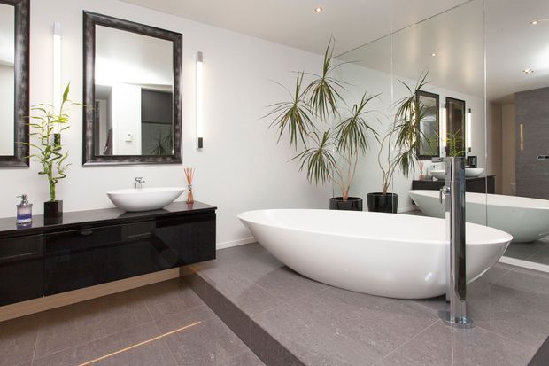 Contemporary bathroom with a freestanding white oval bathtub, wall-mounted black vanity with a white vessel basin, large mirrored wall, tall chrome tapware, grey tiled flooring, and indoor plants. The room is bright and spacious with a refined monochromatic palette.