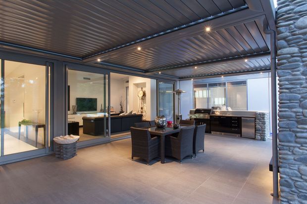 Covered outdoor entertaining area with a dining setting, integrated outdoor kitchen and barbecue, large-format tiled flooring, dark timber-look slatted ceiling, extensive glazing, and distinctive rounded-stone feature walls. Interior living spaces are visible through the glass doors.