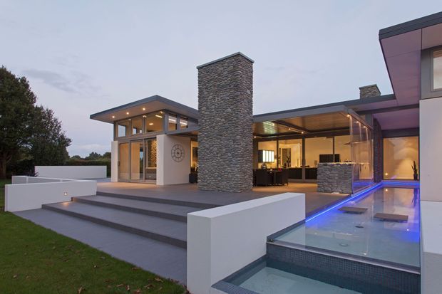Contemporary luxury residence viewed from the landscaped exterior, featuring a flat-roofed composition, extensive floor-to-ceiling glazing, a prominent stacked-stone chimney, broad entry steps, and a raised illuminated swimming pool beside an open-plan interior.