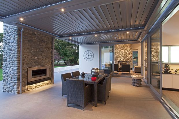 Covered outdoor entertaining area with a dining table and woven chairs, integrated stone fireplace, and a large louvred pergola roof. Glazed sliding doors connect the space to a contemporary interior, with textured stacked-stone walls and warm recessed lighting.