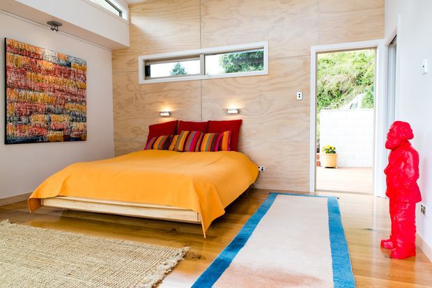 Bright contemporary bedroom with a low timber platform bed dressed in vivid orange bedding and multicoloured striped cushions. The room features pale plywood wall panelling, high horizontal windows, integrated wall lights, timber flooring, and a large blue-bordered rug. An open doorway leads to an exterior terrace or garden, while a bold red sculptural figure provides an accent.