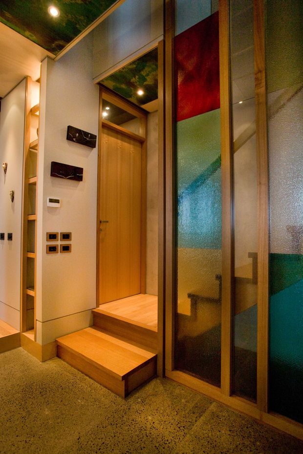 A warmly lit interior entryway or hallway with a short set of timber steps leading to a timber door. Full-height timber-framed textured and coloured glazing forms a prominent partition on the right, while built-in open shelving lines the left wall. The space features polished speckled concrete flooring, pale walls, timber trim, recessed ceiling lights, and colourful translucent glass panels in red, blue, green, and amber.