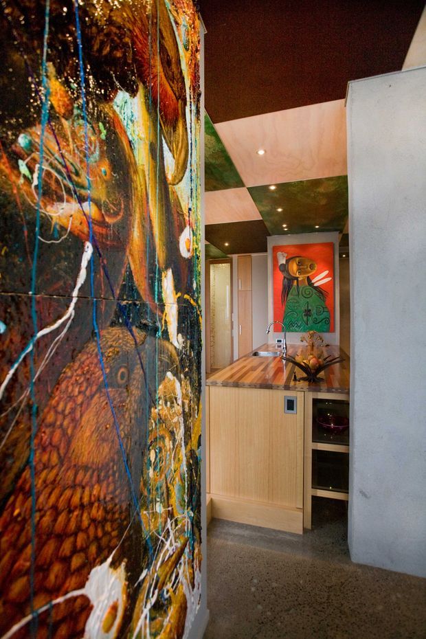 A compact contemporary kitchen viewed through a striking abstract painted feature wall. The kitchen has light timber cabinetry, a timber butcher-block benchtop, integrated sink and tap, open wine storage, and a colourful artwork panel at the rear. Recessed spotlights are set into a geometrically patterned dark and pale ceiling, while polished concrete flooring and a plain grey wall complete the space. The vivid abstract wall and artwork create a bold, eclectic character.