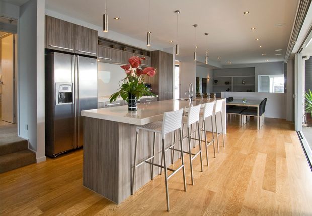 A bright, open-plan contemporary kitchen and dining area with a large central island, white stone benchtop, integrated stainless-steel appliances, dark wood-grain cabinetry, and several white bar stools. Warm timber flooring extends into the dining and living spaces, with recessed and pendant lighting, a pool table in the background, and full-height glazing along the right side.