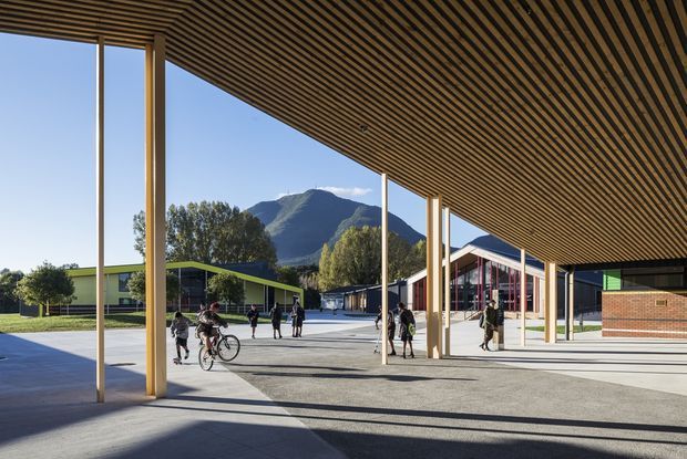 A covered outdoor campus or public building forecourt with a broad timber slatted canopy supported by slender timber columns. Students and bicycles occupy the paved space, with low modern buildings, brickwork, and a mountain landscape visible beyond. The setting is bright and open, with strong linear architectural geometry and a contemporary institutional character.