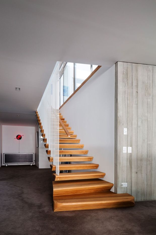 Interior view of a contemporary stairway with open-riser timber stairs, white vertical balustrade rods, and a timber handrail. The staircase rises beside a large glazed opening and a pale grey vertical timber-panelled wall. Dark carpet extends through the surrounding hallway or living area, with a low media console visible in the background.