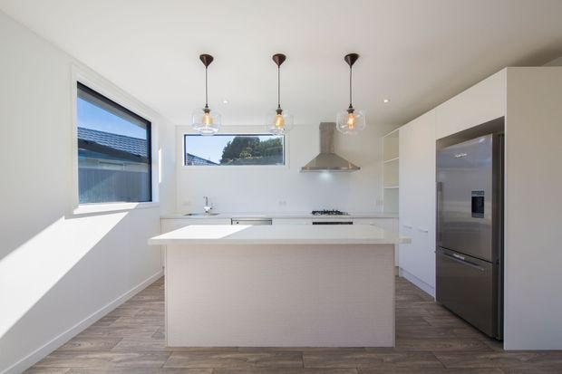 Bright, compact contemporary kitchen with a central island, white handleless cabinetry, pale engineered benchtops, stainless-steel appliances, a stainless rangehood, pendant lighting, and timber-look flooring. Large windows provide strong natural light and views to neighbouring residential buildings.