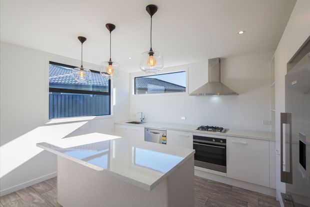 Bright contemporary white kitchen with a central island, integrated cabinetry, stainless-steel appliances, pale engineered stone benchtops, and three pendant lights. Large windows provide strong natural light and overlook a neighbouring tiled roof and fenced exterior.
