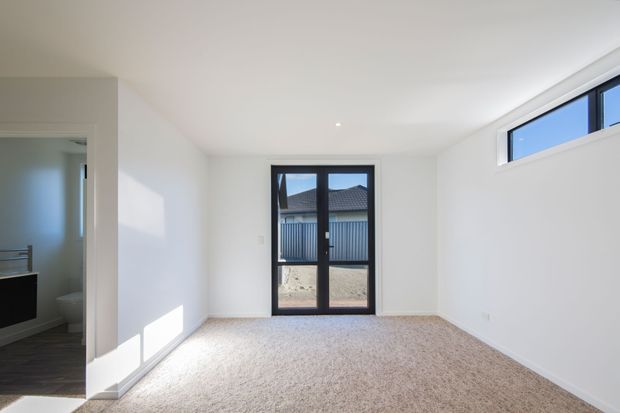 A bright, empty carpeted room with white walls and ceiling, recessed downlight, black-framed glazed door, and a high horizontal window. A partially visible bathroom or adjacent space opens on the left. The room receives strong natural light and has a simple contemporary fit-out.
