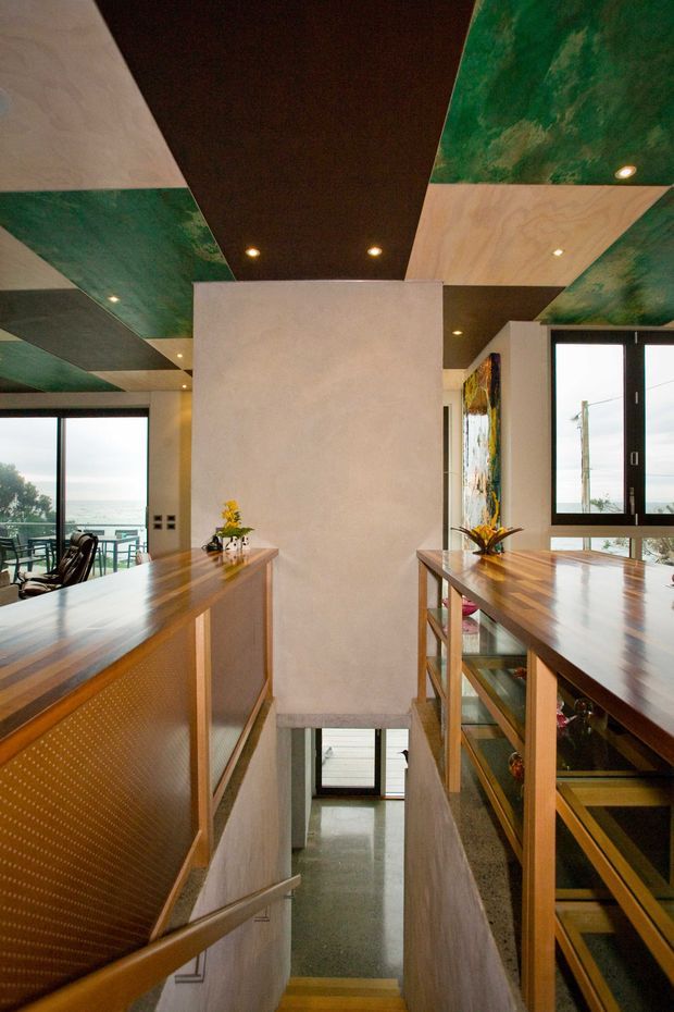 Interior view of a contemporary split-level stairway and landing. Timber-clad balustrades and open shelving flank stairs descending to a lower level, while polished timber counters or ledges frame the landing. The ceiling features a bold geometric patchwork of dark timber, pale timber, and mottled green panels with recessed downlights. Large black-framed windows provide abundant natural light and views outside. Small decorative plants and artwork add colour.