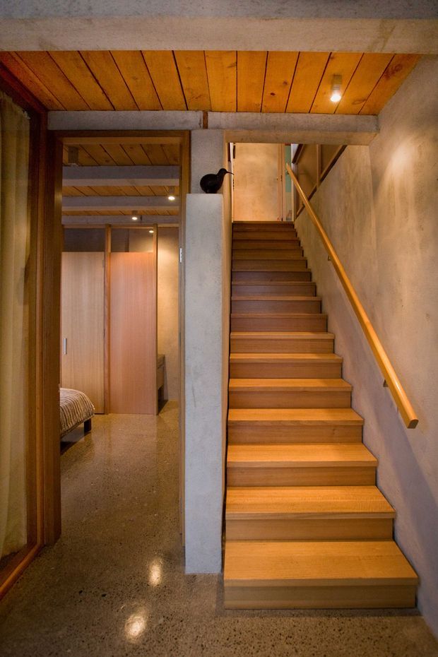 Interior view of a compact stairway rising beside an open bedroom doorway. The staircase has warm timber treads and a timber handrail, with exposed timber ceiling boards, raw concrete beams and column, plastered walls, and a polished terrazzo or concrete floor. Recessed lighting creates a warm, earthy atmosphere.