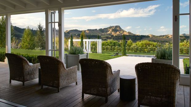 Covered outdoor terrace or sunroom with timber decking, exposed white-painted ceiling beams, and large open glazed doors framing a landscaped garden, pool, vineyard-like planting, and dramatic mountain landscape. Several woven outdoor armchairs and a small round side table create a relaxed seating area.