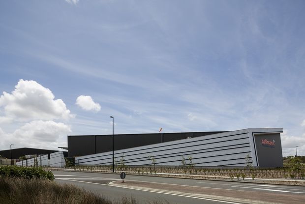 Exterior architectural photograph of a large contemporary commercial or industrial building beside a paved roadway. The building has a long, low profile, dark charcoal cladding, a prominent angled facade with repeated horizontal metal bands, and a dark end wall bearing a small red wordmark. Sparse landscaped planting and a pedestrian path run along the street, beneath a bright blue sky.