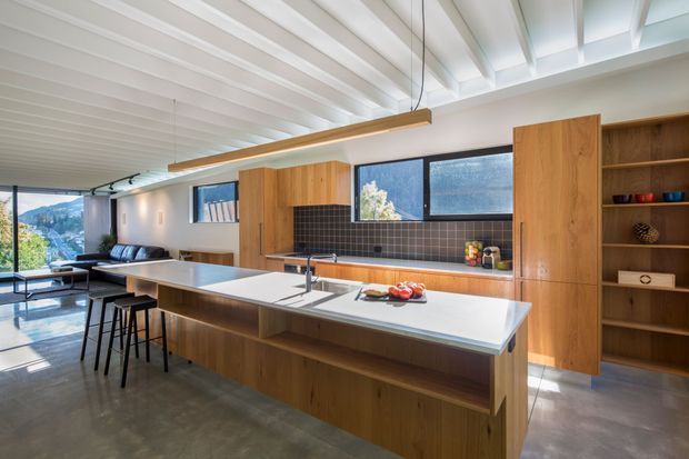 Open-plan contemporary kitchen with a large white island, timber cabinetry, dark tiled splashback, and integrated dining-height seating. The space opens into a living area with expansive glazing and views beyond. A white-painted exposed-beam ceiling and linear pendant lights add strong architectural character.